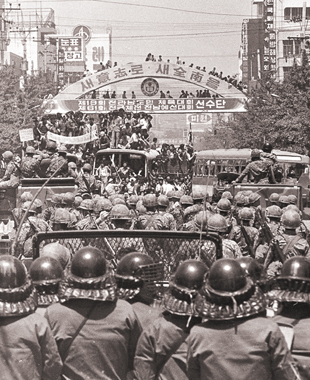 계엄령 철폐를 외치는 시민들이 광주 동구 금남로 가톨릭센터 앞에서 공수부대원 및 경찰과 대치하고 있다. 19회 전라남도민 체육대회 아치가 당시를 알려주고 있다.