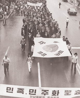 대형태극기를 앞세우고 민족민주화대성회 참석을 위해 교문을 벗어나 금남로로 향하고 있는 전남대학교 교수들, 이들 뒤를 학생들이 따르며 민주주의를 위한 구호를 외치고 있다.